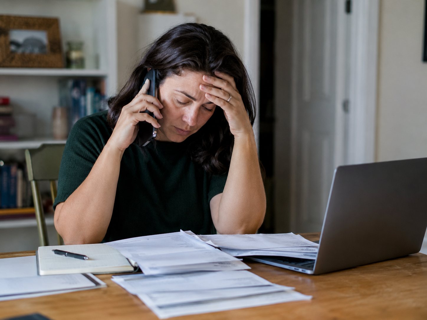 A woman holds a phone to her ear with one hand and her forehead with the other, paperwork and a laptop in front of her