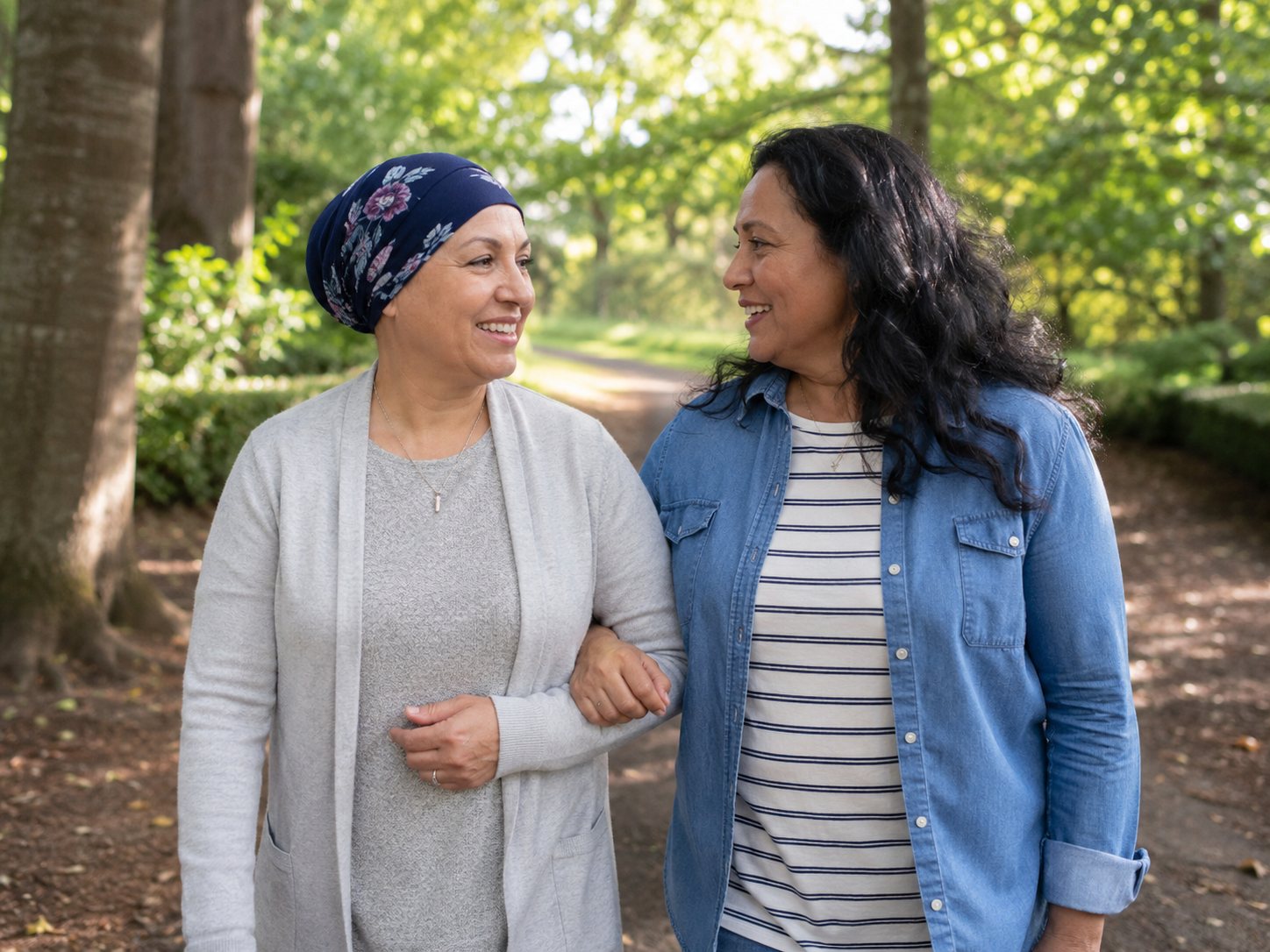Two smiling women link arms while walking a woodland path, one wearing a floral head scarf.