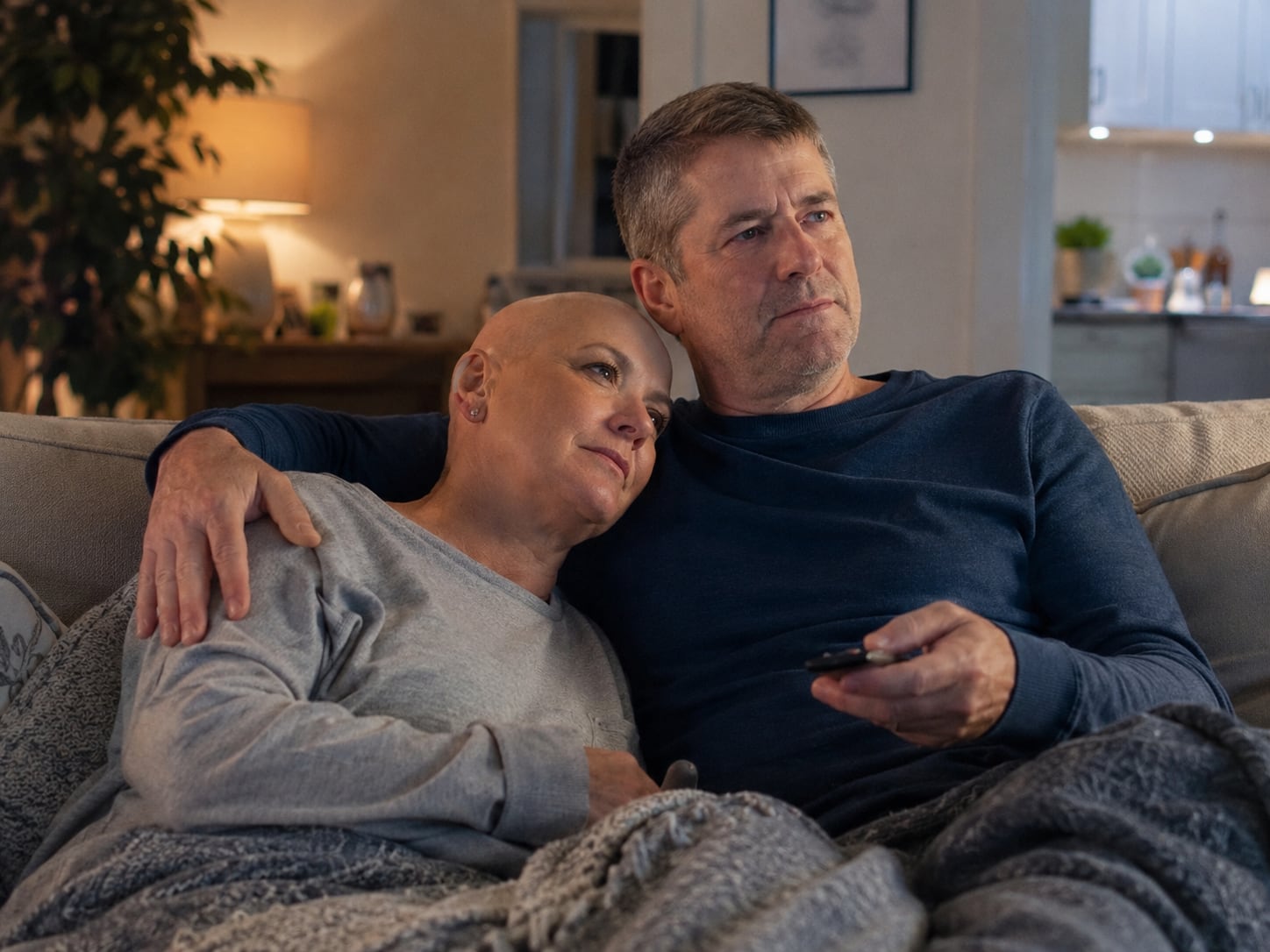 Two men sit on a couch, one resting his head on the other's shoulder in comfort