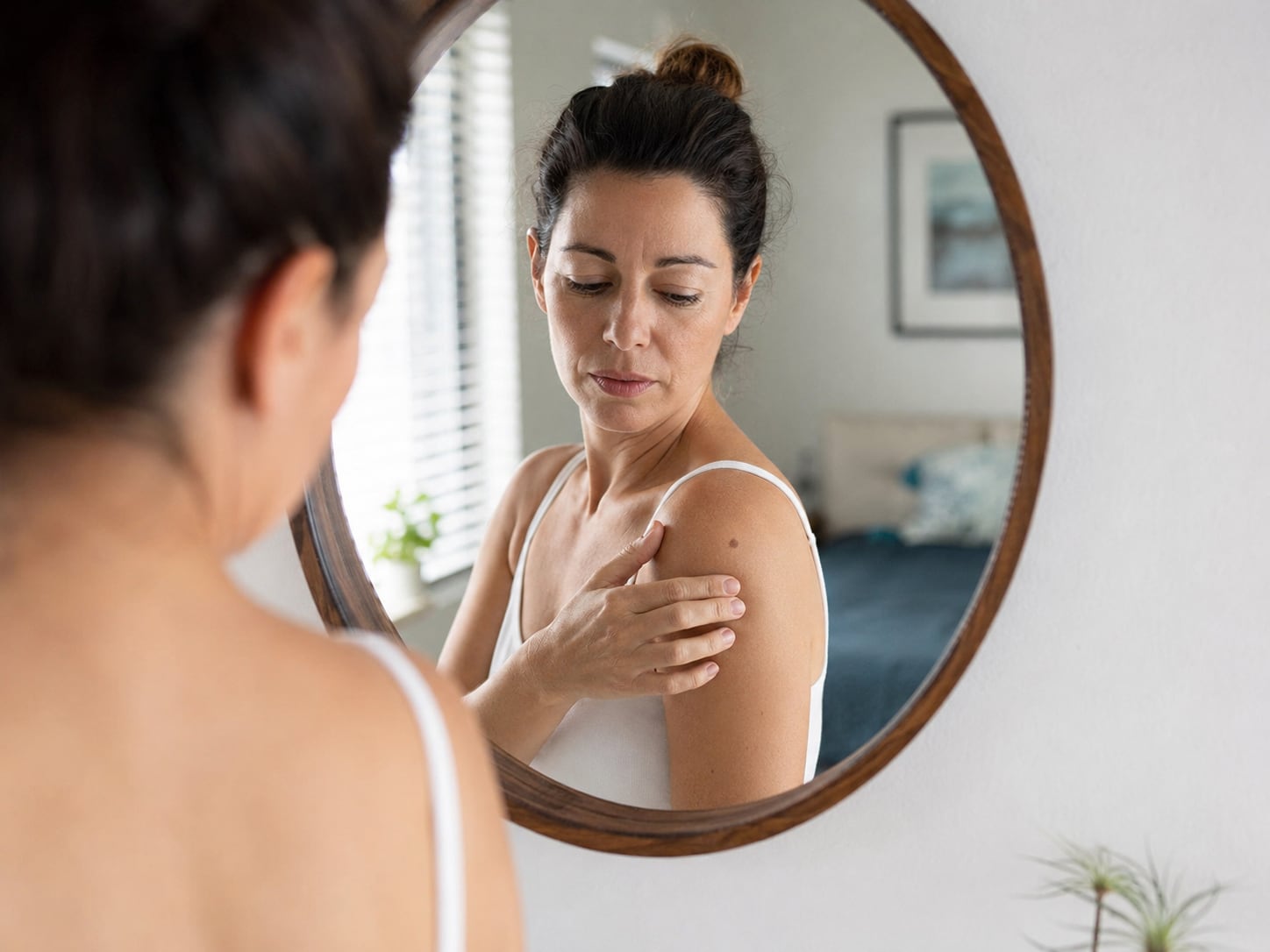 A woman in a mirror examines her bare shoulder and chest, self-exam