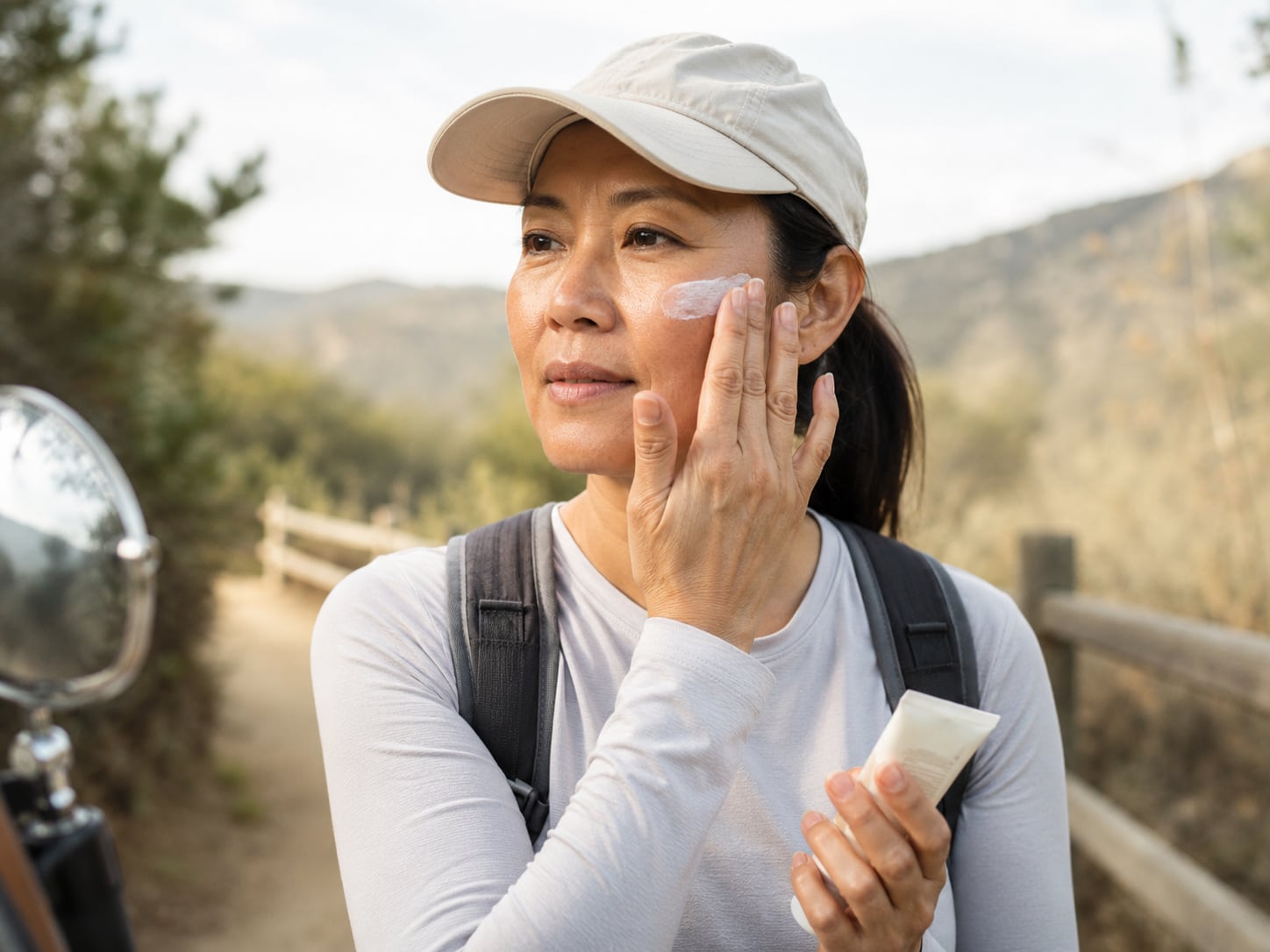 A woman in a cap and backpack applies sunscreen to her face outdoors