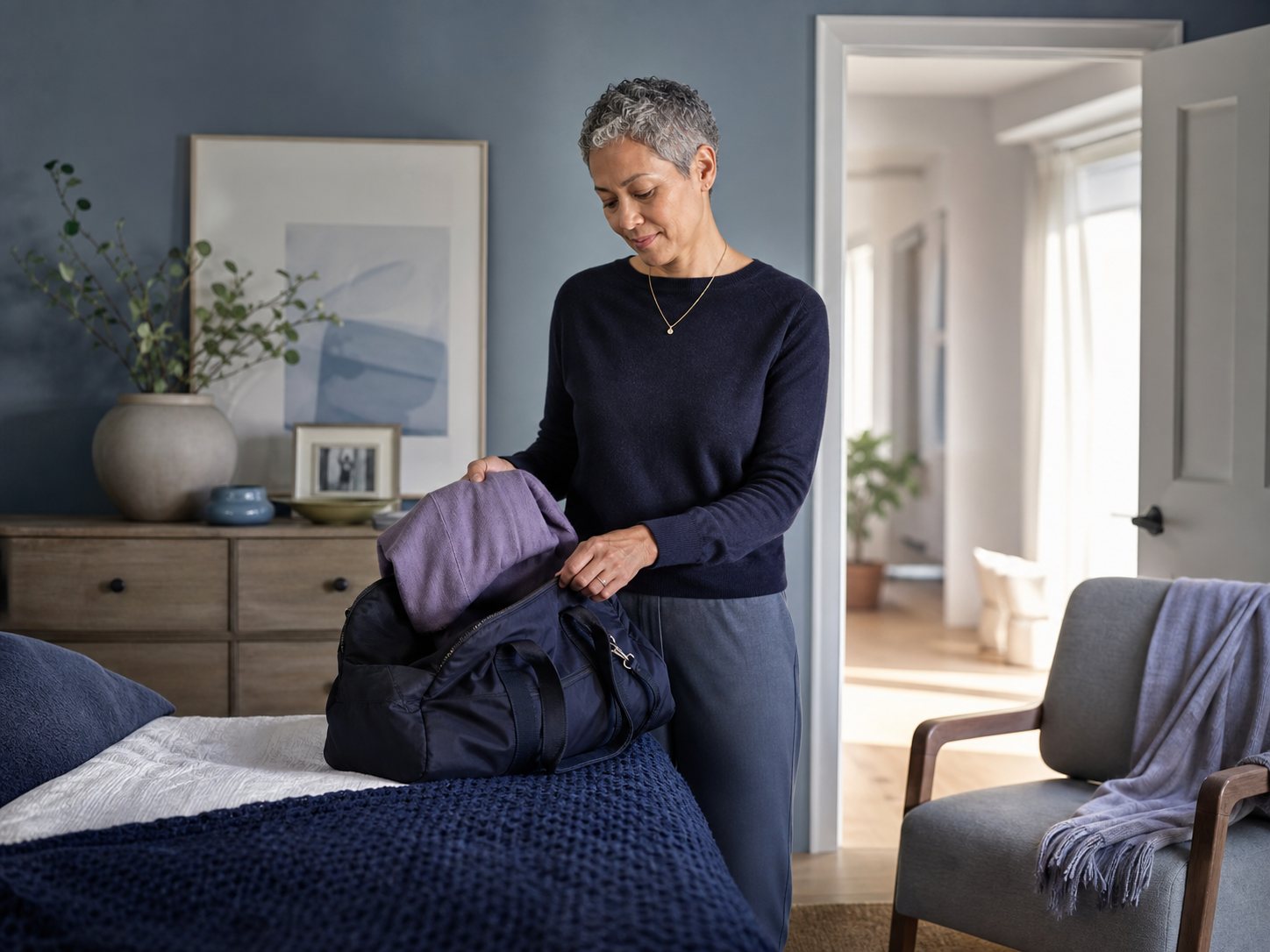 A woman with short hair folds a sweater into a duffel bag on a bed.