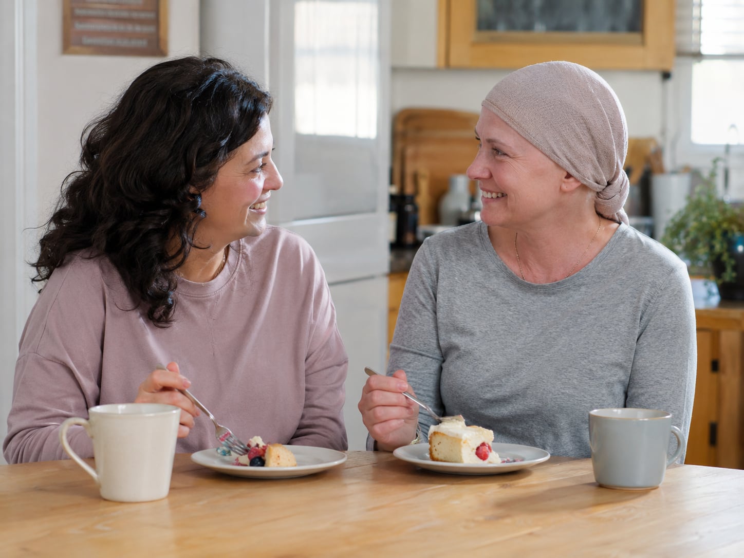 Two women, one wearing a headscarf, sit together smiling with drinks