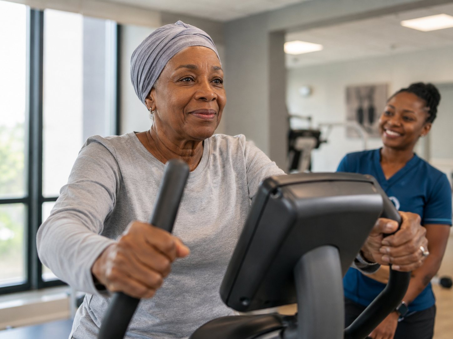 Older woman in a head wrap exercises on a gym machine as a staff member in scrubs looks on.