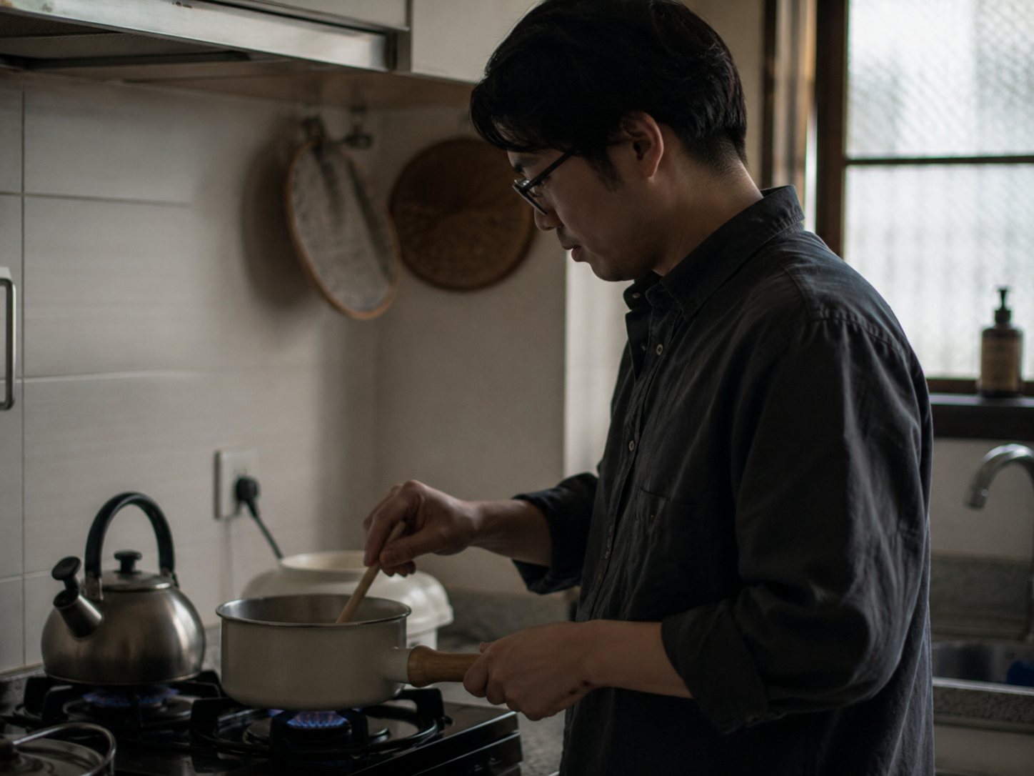 A man stands at a gas stove stirring a small saucepan in a narrow home kitchen