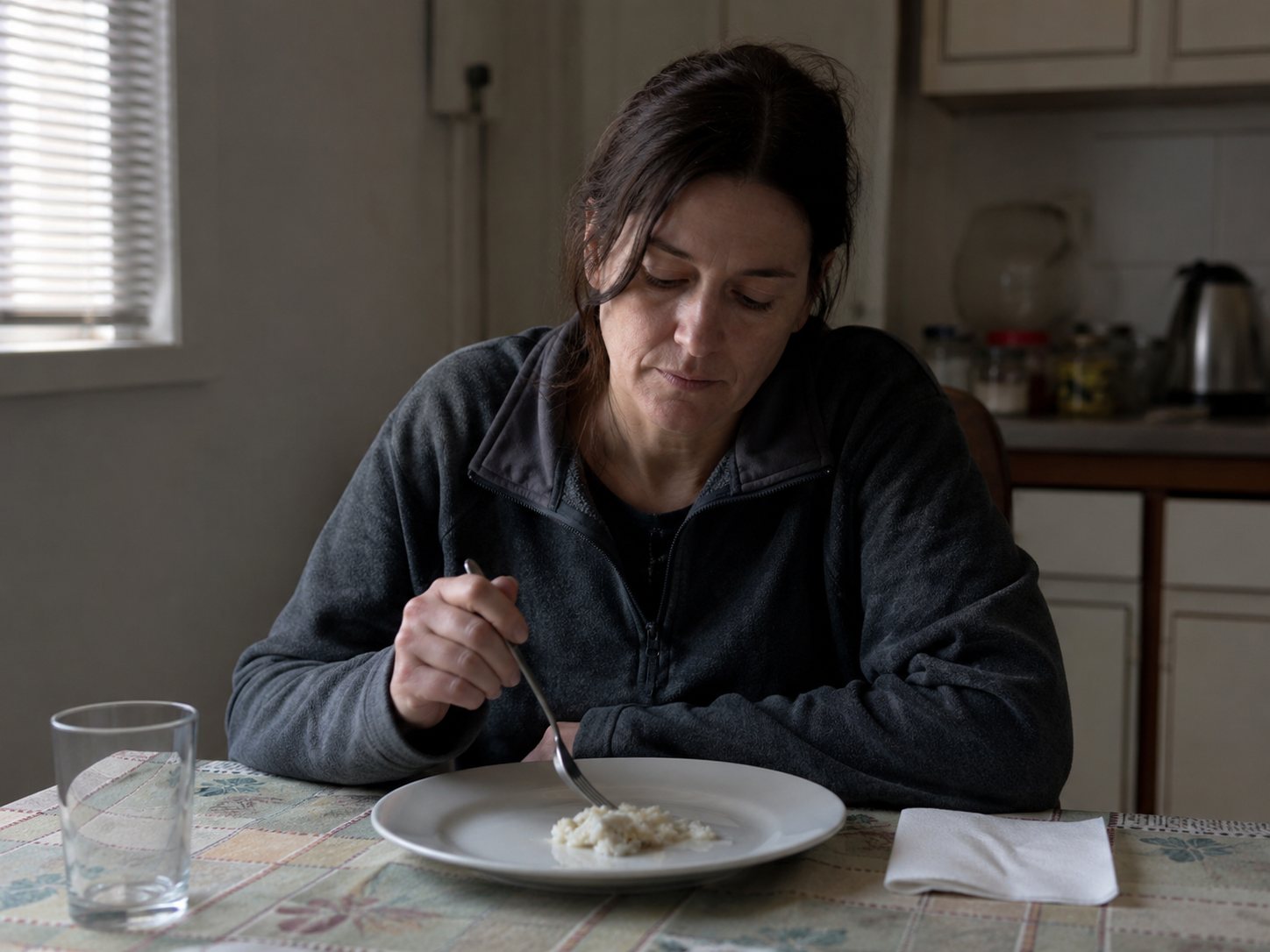 A woman sits alone at a kitchen table looking down at a plate holding only a few spoonfuls of food