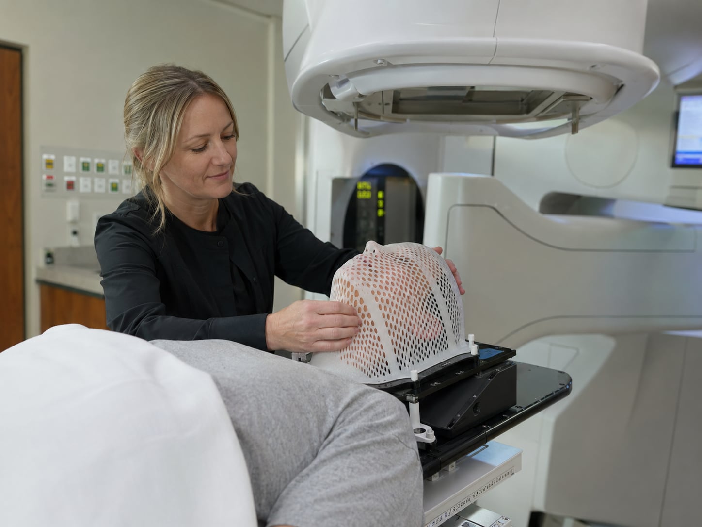 A staff member fits a white perforated thermoplastic mask over the head of a person lying on a radiotherapy treatment couch.