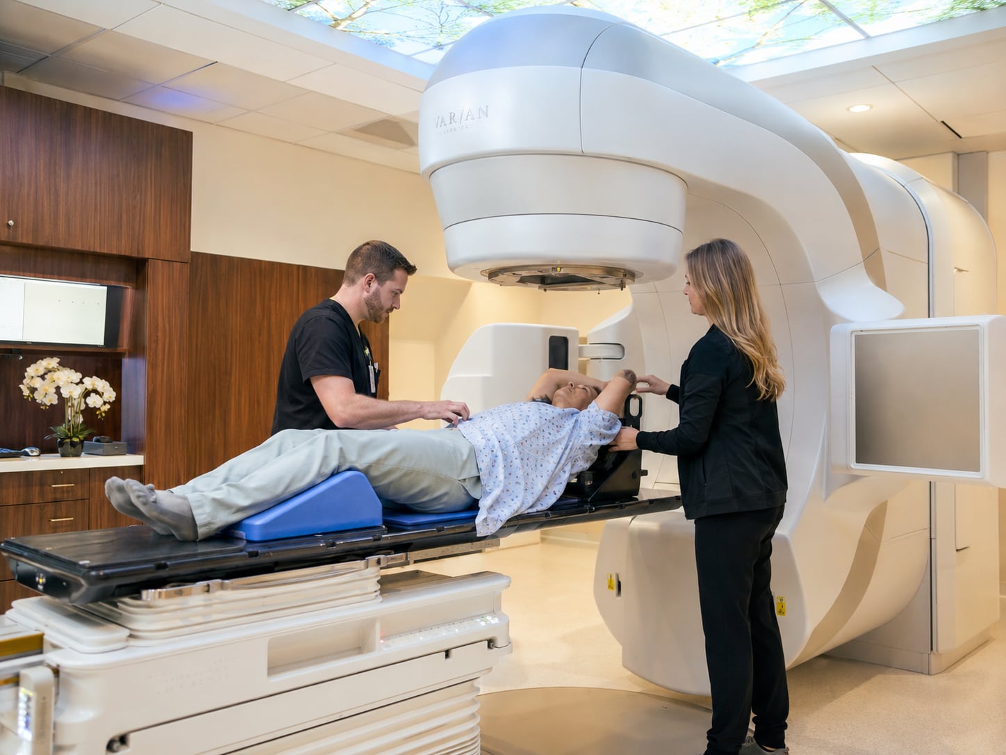 Two staff position a patient in a hospital gown, arms raised overhead, on the couch of a linear accelerator in a radiotherapy room.