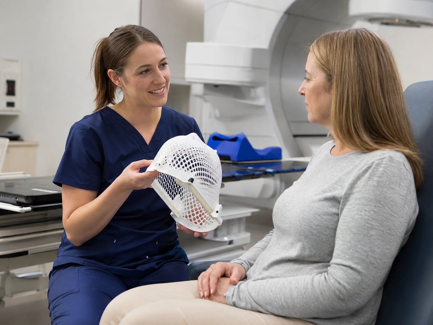 A clinician in navy scrubs holds a white mesh thermoplastic immobilisation mask while talking with a seated woman in front of a linear accelerator.